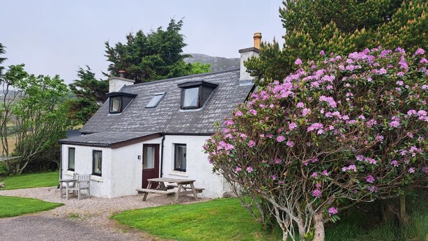 A white cottage with a sloped roof, a chimney, and a small porch with a bench.