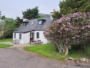 A white cottage with a sloped roof, a chimney, and a small porch with a bench.