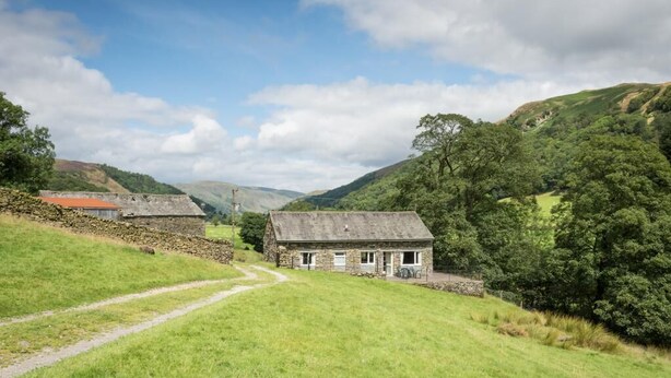 A stone cottage with a grassy yard and a path leading to it, surrounded by hills and trees. - 3 Bed in Kendal (oc-sz466) (Longsleddale)