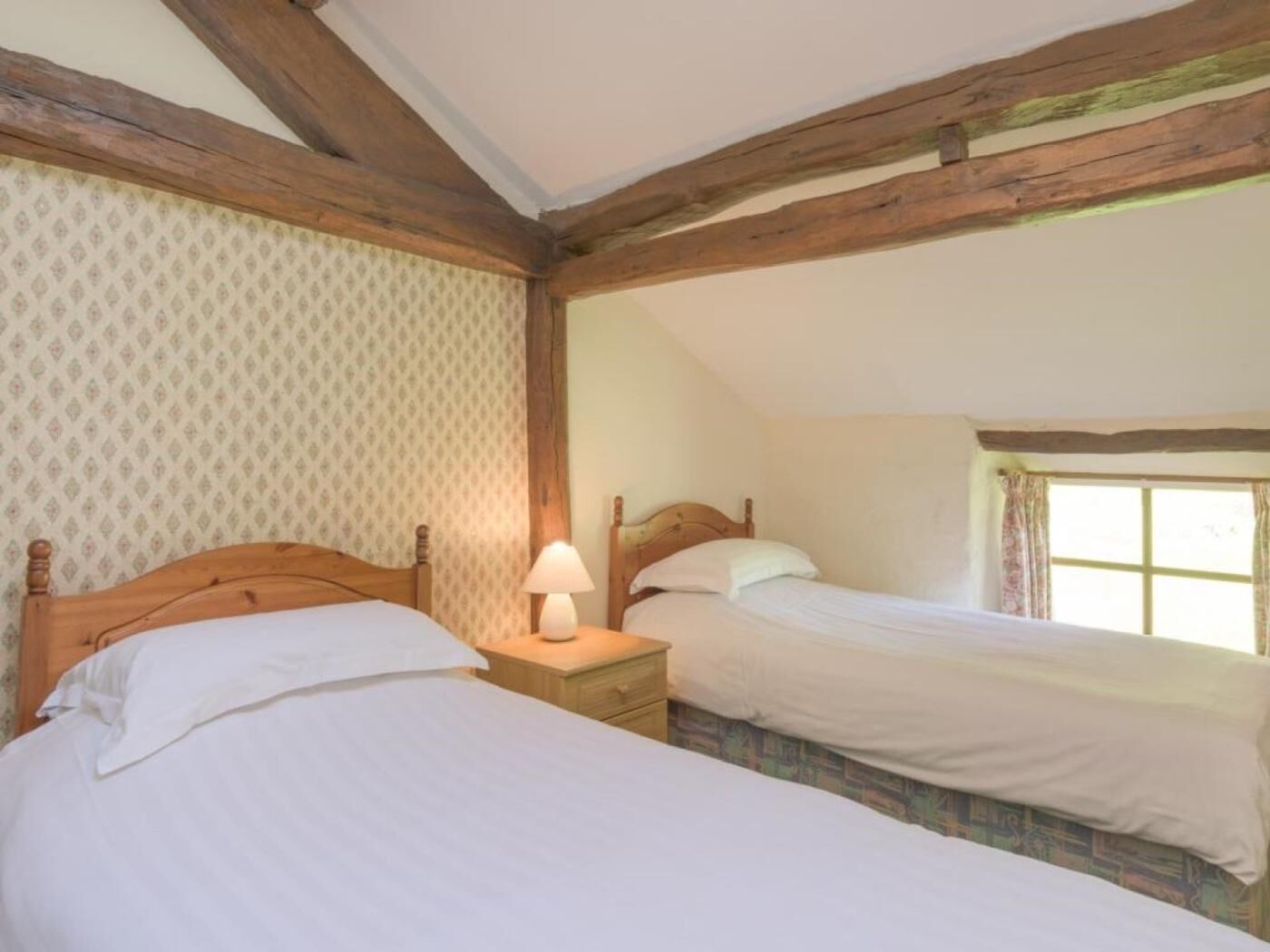 Une chambre avec deux lits simples, des poutres en bois, une fenêtre avec des rideaux et un papier peint à motifs.
