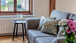A cozy room with a grey sofa, a small table with a glass and bottle, and a window with a view of buildings. - 1 Bed in Bucknell (oc-94661) (Letton)