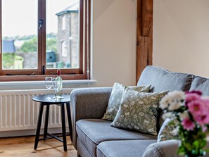 Ein gemütliches Zimmer mit einem grauen Sofa, einem kleinen Tisch mit Glas und Flasche sowie einem Fenster mit Blick auf Gebäude.