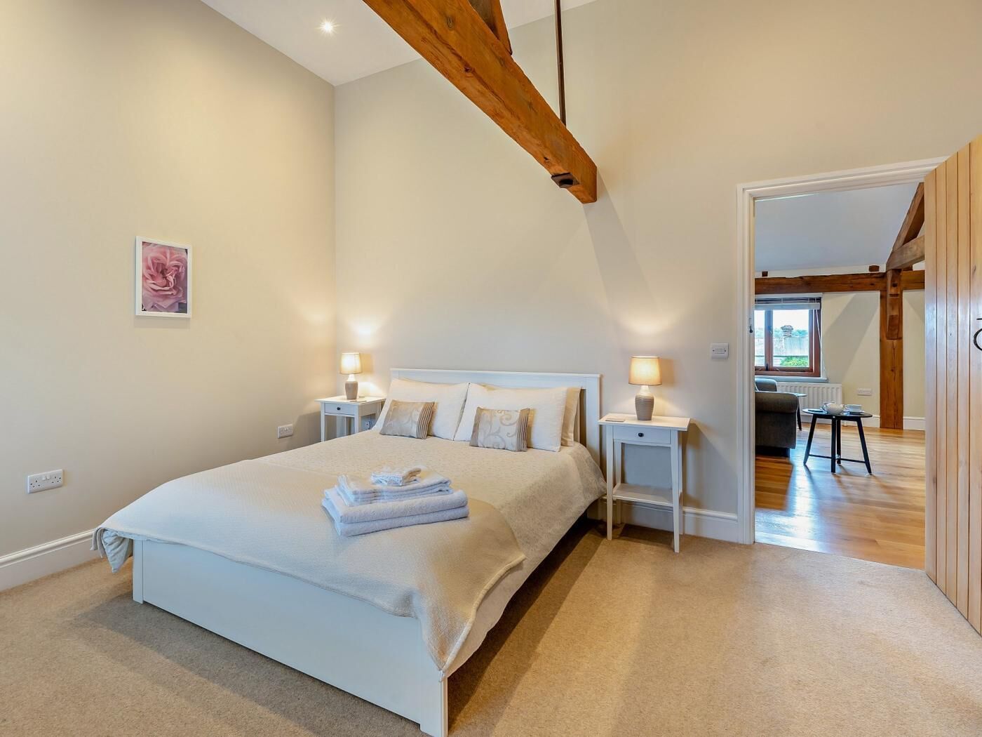 A bedroom with a large bed, bedside tables with lamps, a wooden ceiling beam, and a view into another room.