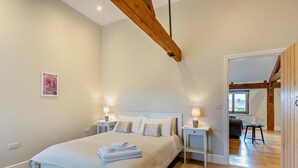 Quarto com cama grande, mesas de cabeceira com abajures, vigas de madeira no teto e vista para outro cômodo.