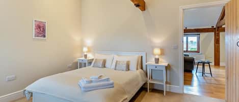 A bedroom with a large bed, bedside tables with lamps, a wooden ceiling beam, and a view into another room.