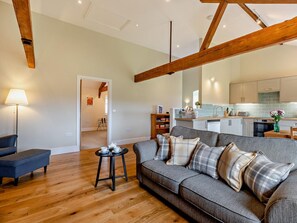 A modern living room with a grey sofa, wooden beams, a small round table with cups, and a kitchen in the background. - 1 Bed in Bucknell (oc-94662) (Letton)