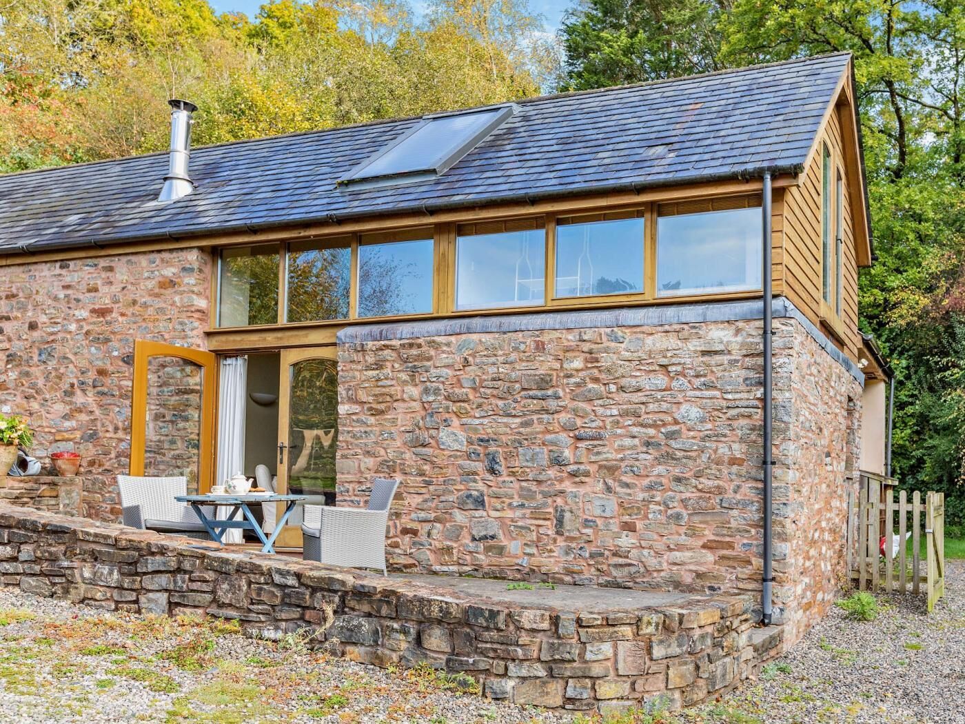 Una casa de piedra con un amplio ventanal, chimenea y un patio con mobiliario exterior.