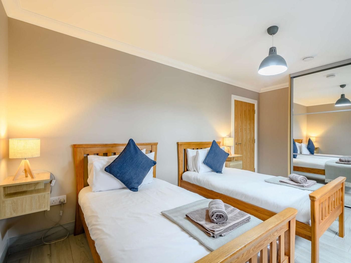 Une chambre d’hôtel avec deux lits, des têtes de lit en bois, une table de chevet avec une lampe, un miroir et un banc en bois.