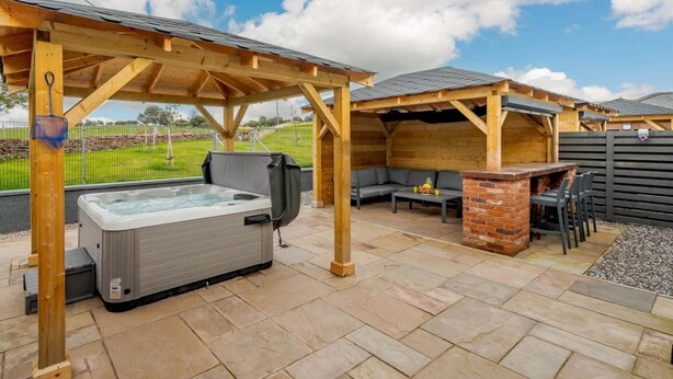 Un bain à remous sous une pergola en bois, un espace barbecue en briques et un coin salon avec vue sur un champ verdoyant.