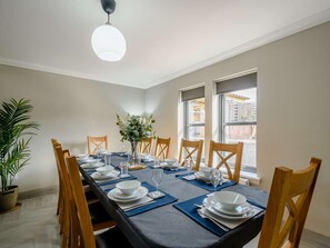 A dining area with a long table set for six, wooden chairs, a vase with flowers, and a view of the city through large windows. - 4 Bed in Alexandria (oc-88759) (Balloch)