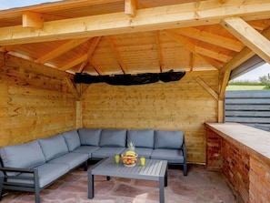 A covered outdoor seating area with a built-in bench and table, featuring a brick wall and wooden beams. - 4 Bed in Alexandria (oc-88759) (Balloch)