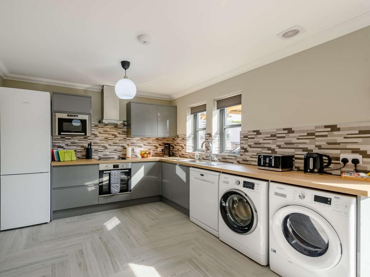 A compact kitchen with a microwave, oven, and washing machine, featuring a herringbone tile backsplash and a wooden countertop.