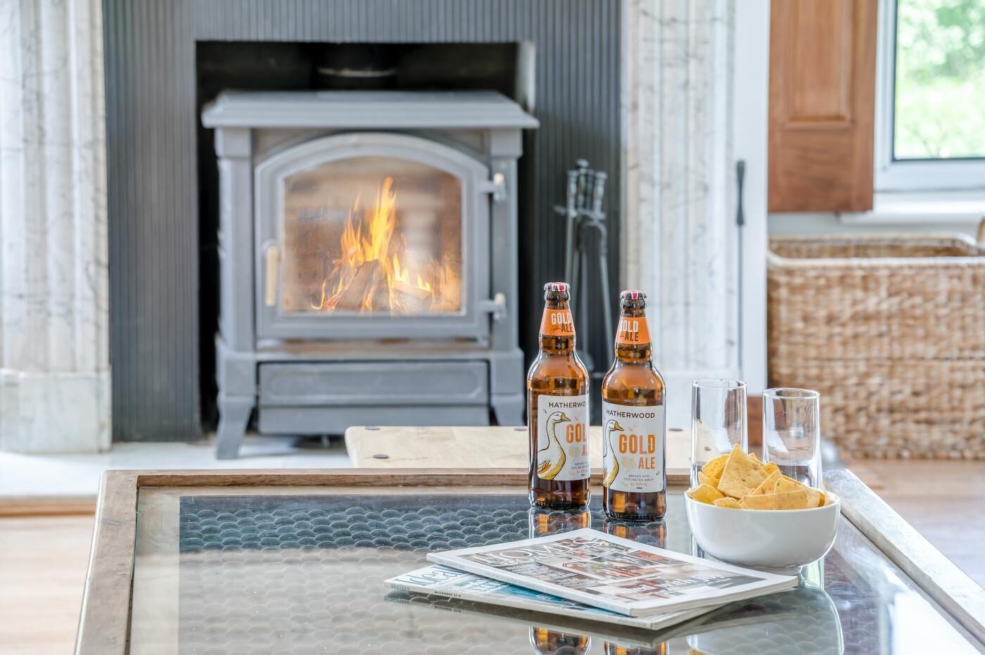 A cozy room with a fireplace, a glass table with a bowl of chips, two bottles of Hatherwood Gold Ale, and a magazine.