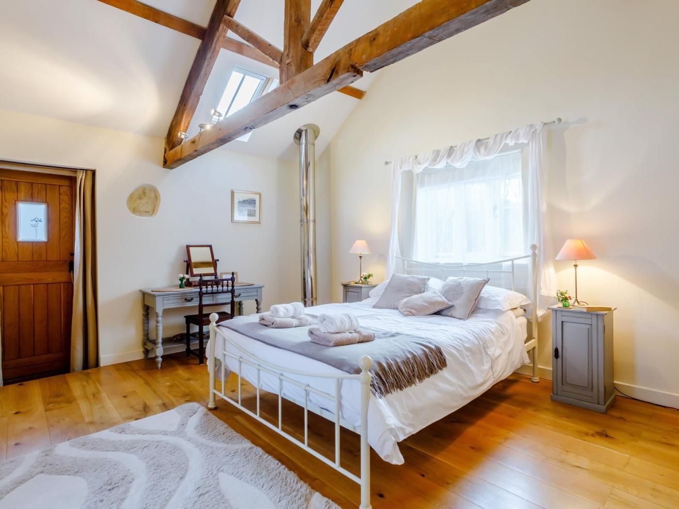 Een slaapkamer met een bed, nachtkastjes, een bureau, een stoel, een raam met gordijnen, een houten plafond en een deur.