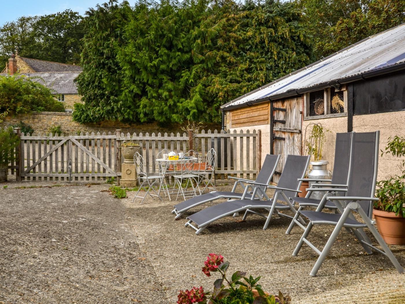 A gravel patio with lounge chairs and a table, surrounded by a wooden fence and a building with a metal roof.