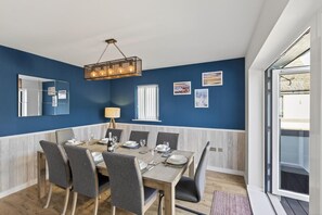 A dining area with a table set for six, a hanging light fixture, and a view through a glass door. - 4 Bed in Bideford (oc-94726) (Westward Ho!)