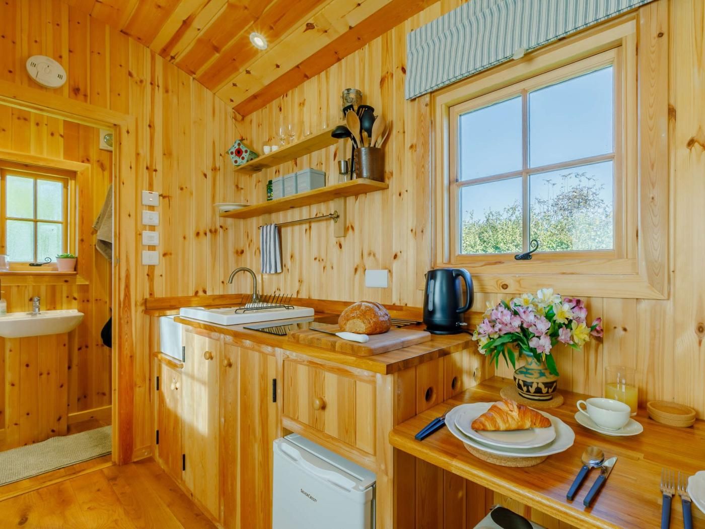 A wooden cabin interior with a kitchen area, a sink, a refrigerator, a window with a view, and a dining table set with plates, a cup, and a vase of flowers.