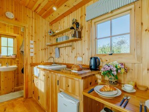 A wooden cabin interior with a kitchen area, a sink, a refrigerator, a window with a view, and a dining table set with plates, a cup, and a vase of flowers. - 1 Bed in Taunton (oc-89989) (Churchstanton)