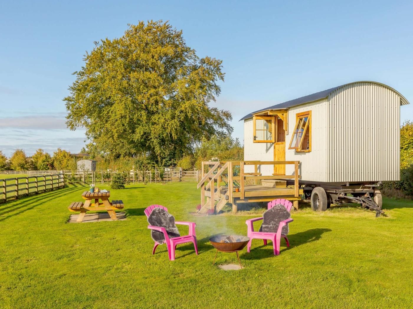 A mobile home with a deck, two chairs, a fire pit, and a large tree in the background.