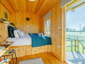 A wooden cabin room with a bed, a small table, and a view of a green field. - 1 Bed in Taunton (oc-89989) (Churchstanton)
