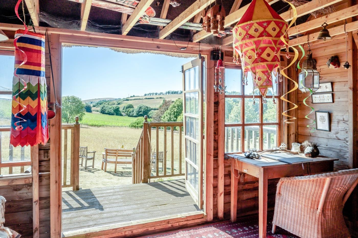 A wooden cabin with a large open door leading to a deck, a table with a chair, and a hanging decorative item.