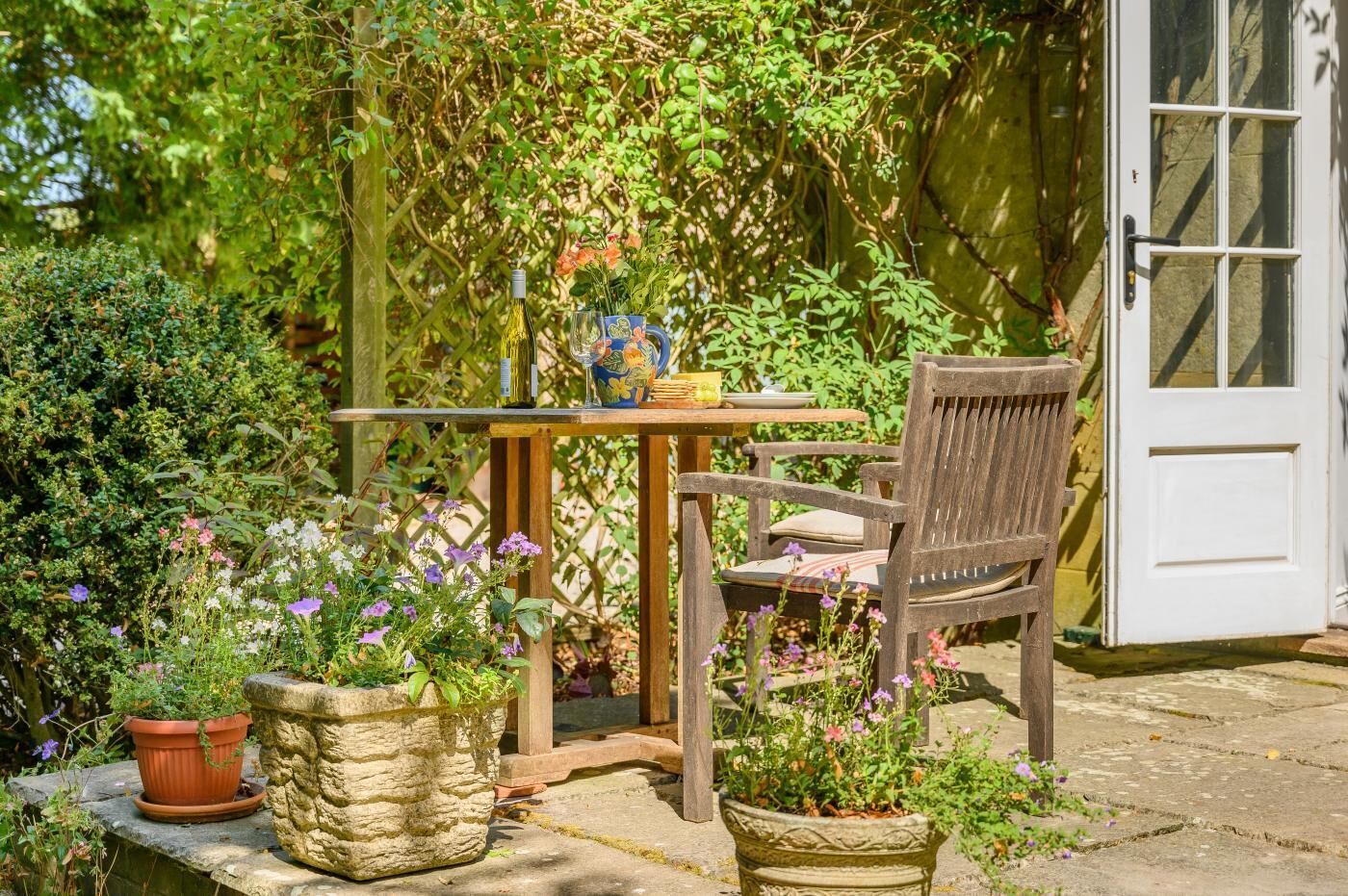 Une table en bois avec une bouteille et un objet décoratif, une chaise en bois et des plantes en pot, le tout dans un décor de jardin.