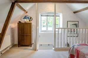 Ein Zimmer mit schrägen Wänden, einem Holzschrank, einem Fenster mit Blick ins Grüne und einem weißen Geländer.