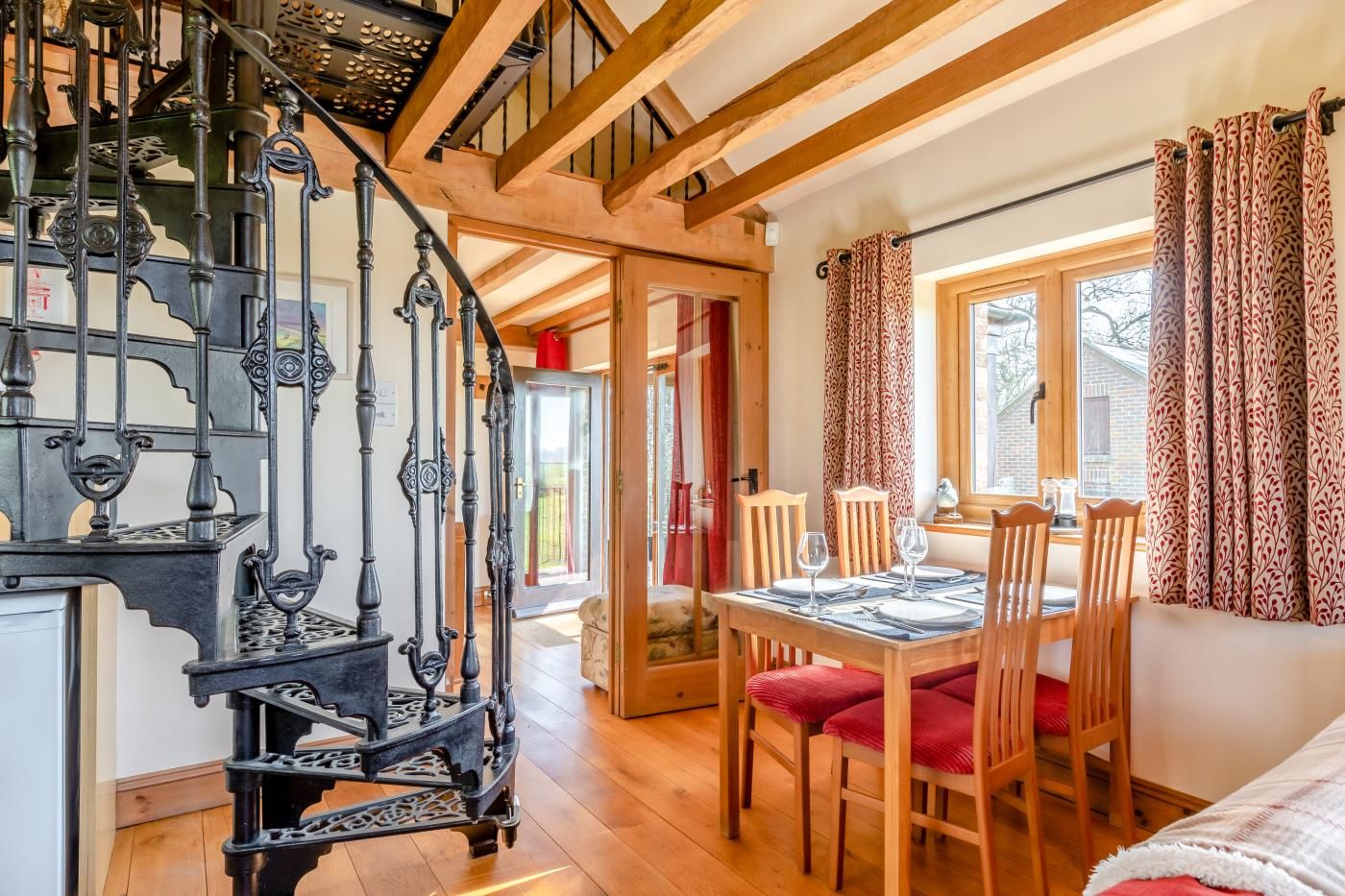 A dining area with a wooden table and chairs, a staircase with ornate metal railings, and a view through a window with patterned curtains.