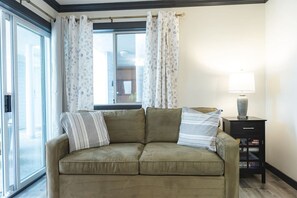 A living room with a sofa, two pillows, a lamp, a small table, and a window with curtains. - Ocean Lookout - Resort Stay with Pool & Hot Tub (Nanoose Bay)