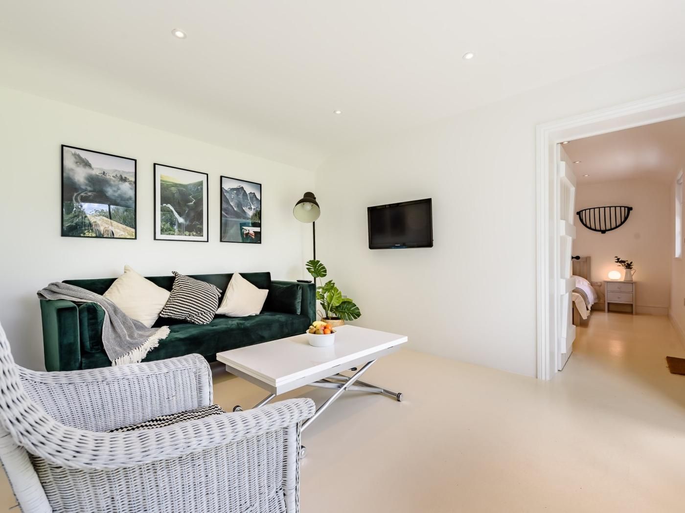 A modern living room with a green sofa, white coffee table, and wicker chairs. There are framed pictures on the wall, a flat-screen TV, and a lamp.