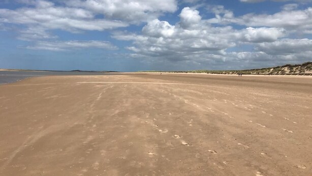 En bred sandstrand med fodaftryk, en klar himmel og fjerne klitter.