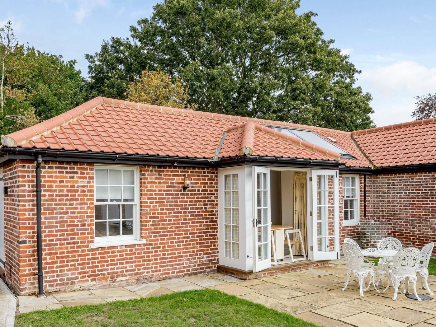 A brick house with a tiled roof, a patio with white outdoor furniture, and a large tree in the background.