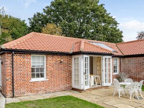 A brick house with a tiled roof, a patio with white outdoor furniture, and a large tree in the background. - 1 Bed in Manningtree (oc-78388) (Lawford)