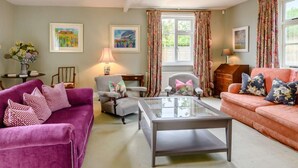 A living room with a purple sofa, a grey armchair, a glass coffee table, and a pink sofa. There are two windows with floral curtains, a lamp on a side table, and framed artwork on the walls. - 5 Bed in Hunstanton (oc-kt185) (Sedgeford)