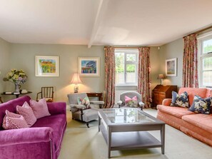 A living room with a purple sofa, a grey armchair, a glass coffee table, and a pink sofa. There are two windows with floral curtains, a lamp on a side table, and framed artwork on the walls. - 5 Bed in Hunstanton (oc-kt185) (Sedgeford)