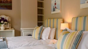 A hotel room with two beds, a fireplace, a nightstand with a vase of flowers, and artwork on the walls. - 5 Bed in Hunstanton (oc-kt185) (Sedgeford)