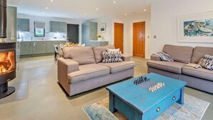 A modern living room with a fireplace, two sofas, a coffee table, and a kitchen in the background. - 2 Bed in Holsworthy (oc-50598) (Sessacott)