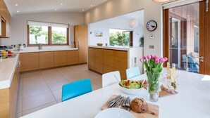 Een moderne keuken met houten kastjes, een eethoek met een witte tafel en uitzicht op de omgeving door grote ramen.