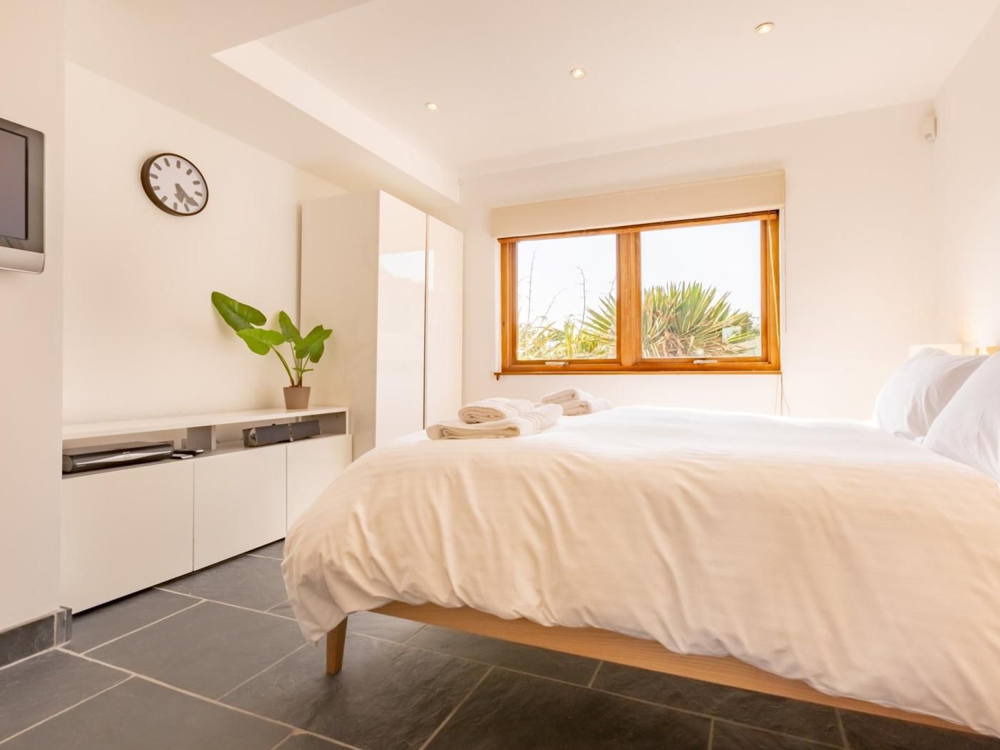 A bedroom with a bed, a potted plant, a clock, and a window with a view.