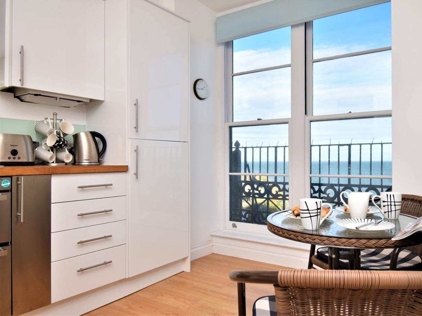 Eine Küche mit weißen Schränken, einem Edelstahl-Wasserkocher und einer Kaffeemaschine. Ein Essbereich mit einem Korbstuhl und einem Tisch für zwei Personen. Ein großes Fenster bietet Meerblick und ein Balkon mit Geländer.