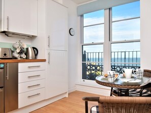 A kitchen with white cabinets, a stainless steel kettle, and a coffee machine. There's a dining area with a wicker chair and a table set for two. A large window offers a view of the sea and a balcony with a railing. - 1 Bed in Ilfracombe (oc-90074) (Ilfracombe)