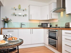A modern kitchen with white cabinets, a microwave, an oven, and a glass shelf with decorative items. - 1 Bed in Ilfracombe (oc-90074) (Ilfracombe)