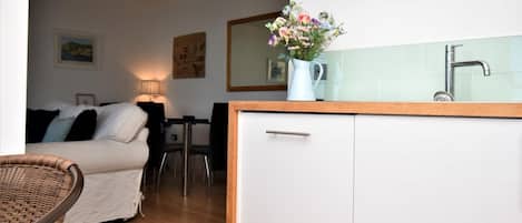 Una cocina compacta con encimera blanca, un lavamanos y un florero con flores. Al fondo, hay una zona de comedor con una mesa y sillas.