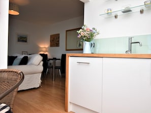 Een compacte keuken met een wit aanrecht, een spoelbak en een vaas met bloemen. Op de achtergrond is een eethoek met een tafel en stoelen te zien.