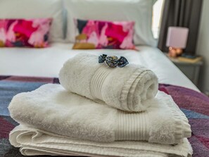 Ein ordentlich bezogenes Bett mit einem weißen Handtuch und einem zusammengerollten Handtuch darauf, einem farbenfrohen Kissen sowie einem Nachttisch mit einer Lampe.