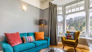 Ein Wohnzimmer mit einem türkisfarbenen Sofa, einem runden Couchtisch, einem gelben Sessel und einem großen Fenster mit Blick auf Bäume und Gebäude.