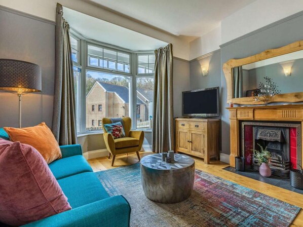 Ein Wohnzimmer mit einer Couch, Sesseln, einem Couchtisch, Kamin, Fernseher und großen Fenstern.