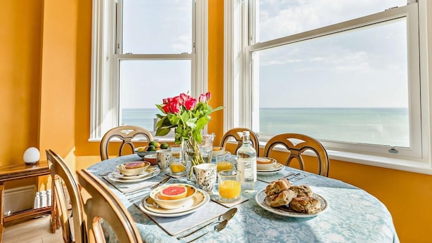 Ein Esszimmer mit gedecktem Tisch, Geschirr, Gläsern und einer Blumenvase, mit Blick auf den Ozean durch große Fenster.