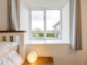 A bedroom with a bed, a bedside table with a lamp, and a window with a view of a house and greenery. - 3 Bed in Ilfracombe (oc-47348) (Berrynarbor)