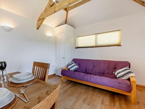 A room with a wooden dining table set for two, a purple sofa, and a window with blinds. - 1 Bed in Launceston (oc-49662) (Saint Clether)